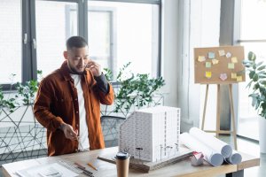 asian architect holding pencil while talking on smartphone near house models on desk