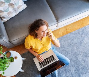 Beautiful young happy female woman in yellow shirt works on laptop at home and talking mobile phone in living room sitting on floor