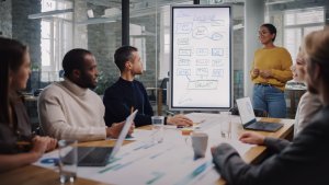 Project Manager Makes a Presentation for a Young Diverse Creative Team in Meeting Room in an Agency. Colleagues Sit Behind Conference Table and Discuss Business Development, User Interface and Design.