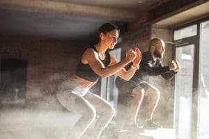 Fit caucasian couple doing squats on boxes in a crossfit style gym, well lit by bright sunlight.