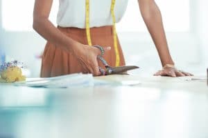 Close-up of professional tailor cutting cloth fabric with scissors while sewing custom made clothes at the table in workshop