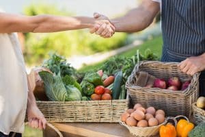 Deux hommes, dont on ne voit que les mains qui se serrent au-dessus d'un étal de légumes frais symbolisant une rencontre autour de la bio.