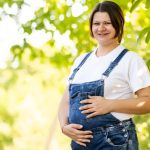 Une femme brune enceinte et souriante, habillée en salopette, au milieu d'un verger