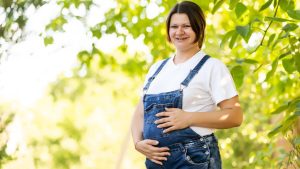 Une femme brune enceinte et souriante, habillée en salopette, au milieu d'un verger