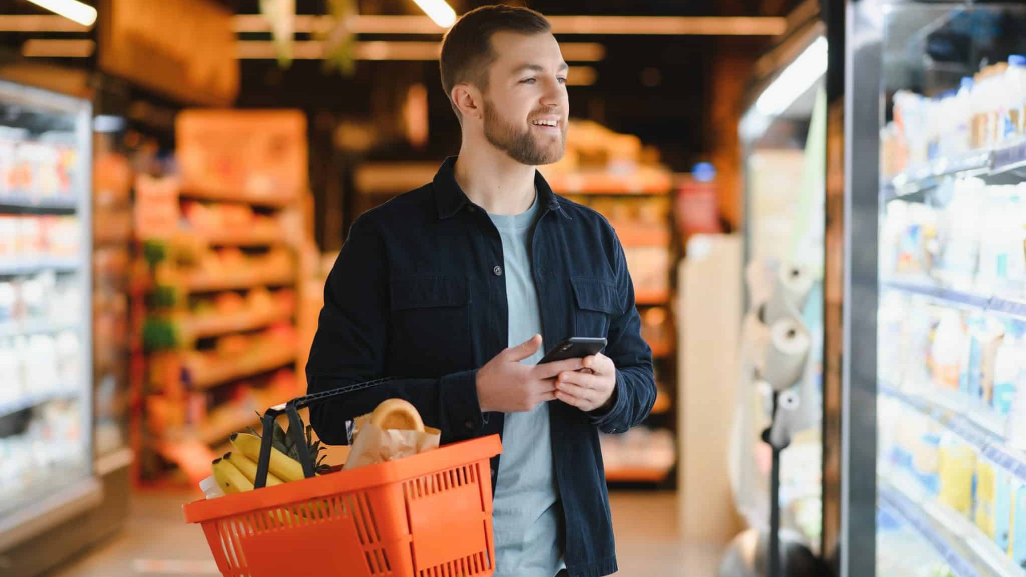 Un homme, seul, au milieu des rayons de produits réfrigérés, porte son smartphone et son panier et sourit en regardant ce qu'il va choisir.