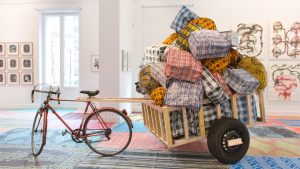 Une œuvre de Barthélémy Toguo. Il s'agit d'un vélo stationné qui a une remorque remplie de nombreux sacs.