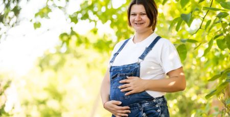 Une femme brune enceinte et souriante, habillée en salopette, au milieu d'un verger