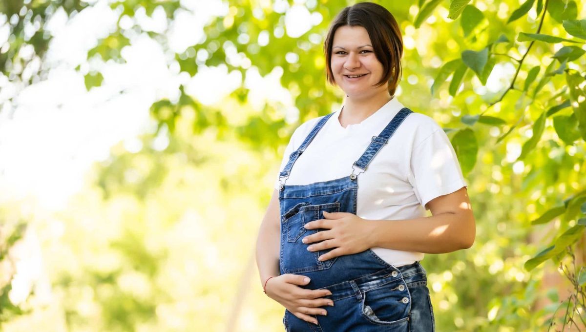 Une femme brune enceinte et souriante, habillée en salopette, au milieu d'un verger