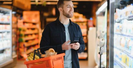 Un homme, seul, au milieu des rayons de produits réfrigérés, porte son smartphone et son panier et sourit en regardant ce qu'il va choisir.