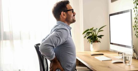 Un homme brun avec des lunettes est assis à un bureau. Il se tient le bas du dos avec ses deux mains car il souffre