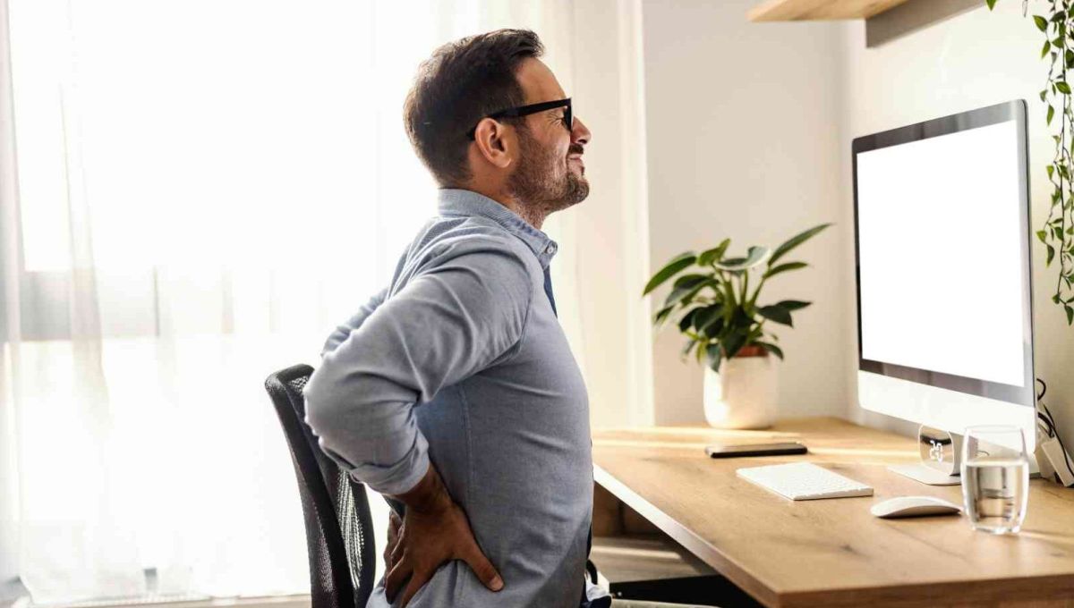 Un homme brun avec des lunettes est assis à un bureau. Il se tient le bas du dos avec ses deux mains car il souffre