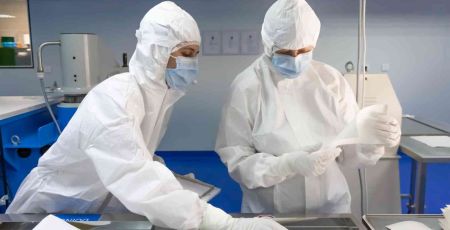 Deux personnes portent une combinaison blanche qui couvre tout leur corps ainsi que la t^te. Elle porte un asque sur le visage. Elles sont dans un laboratoire et regardent une feuille de papier.