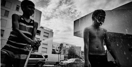 Photo de Loïs Mugen donné au collectif Bienvenue pour une expo-vente au profit de SOS Méditerranée. On voit deux jeuns garçon en bas d'un immeuble jouer avc un tuyau d'arrosage