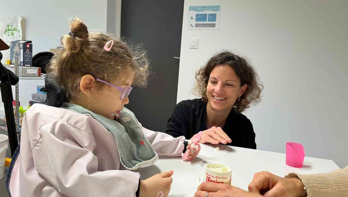 Une petite fille mange un yaourt, encouragée par ses thérapeutes