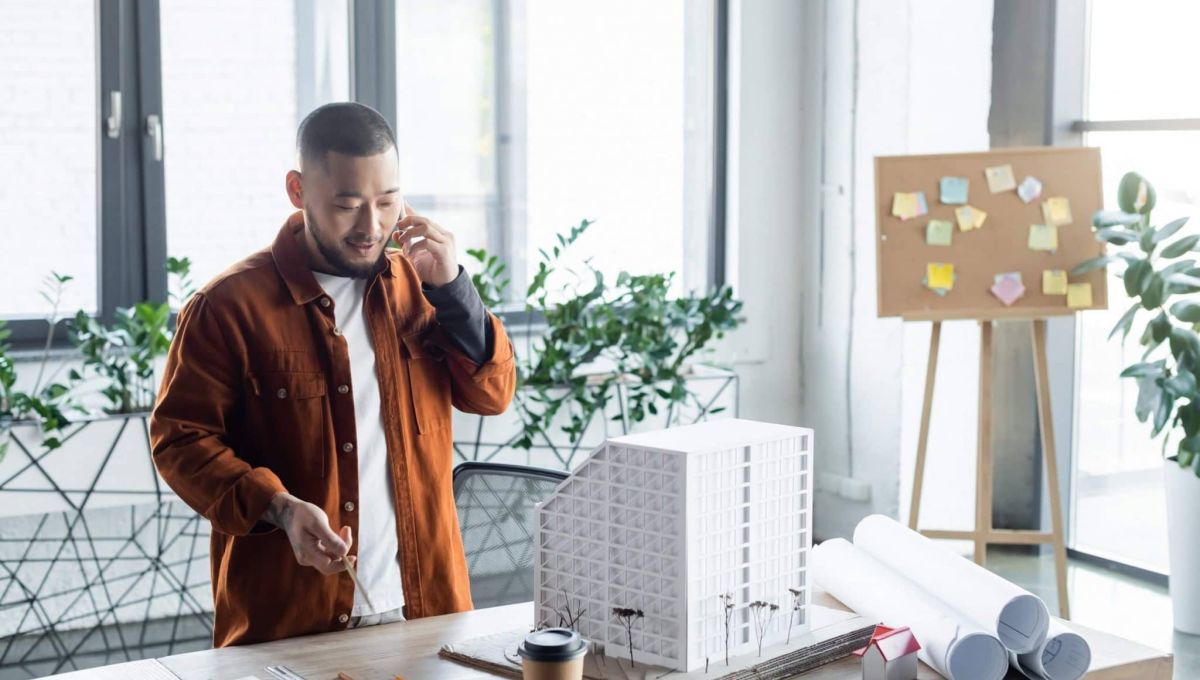 asian architect holding pencil while talking on smartphone near house models on desk