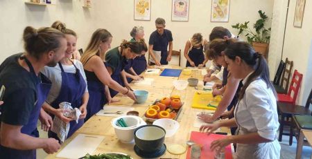 Un atelier culinaire est organisé. Il y a une dizaine de participants autour d'une table. Il porte un tablier bleu