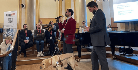 Thibaut de Martimprey chargé de projet du campus Louis Braille est en train de parler au micro. Son chien-guide se teint à ses côtés. Plusieurs personnes sont autour de lui.