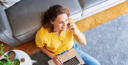 Beautiful young happy female woman in yellow shirt works on laptop at home and talking mobile phone in living room sitting on floor