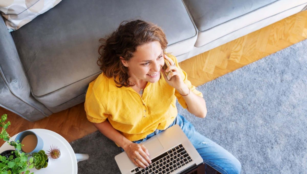 Beautiful young happy female woman in yellow shirt works on laptop at home and talking mobile phone in living room sitting on floor
