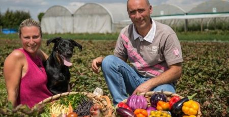 De gauche à droite, une femme, un chien, un homme accroupis au milieu d'un champ et devant leur production de légumes mêlant de nombreuses couleurs.