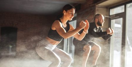 Fit caucasian couple doing squats on boxes in a crossfit style gym, well lit by bright sunlight.