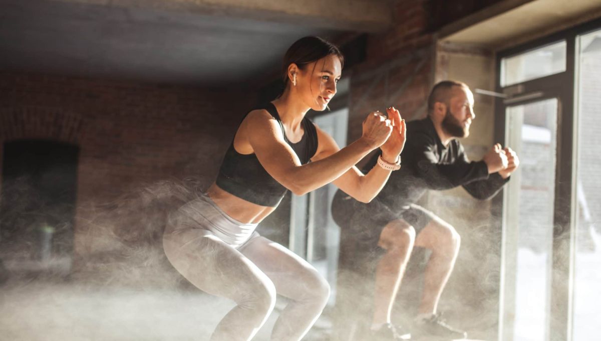 Fit caucasian couple doing squats on boxes in a crossfit style gym, well lit by bright sunlight.