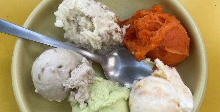 Cinq boules de glace aux couleurs variées sont présentées dans un petit bol beige clair, avec une cuillère en métal posée dessus.