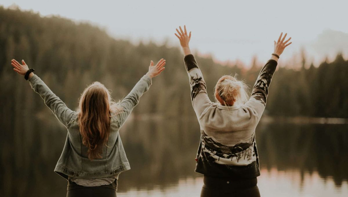 Deux femmes les bras en l'air font face à un lac et expriment leur bonheur
