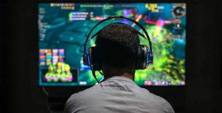 Une personne est de dos. Elle joue à un jeu vidéo sur ordinateur. Elle a un casque sur les oreilles.